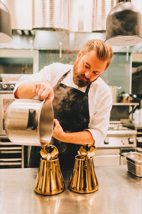 Kock arbetar koncentrerat med en kastrull i ett ljust restaurangkök.
